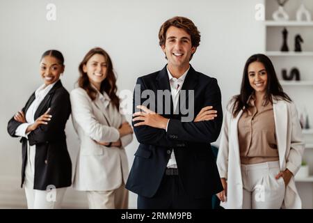 Positiver Geschäftsmann, der mit gefalteten Armen vor seinen multirassischen weiblichen Angestellten steht und im Amt posiert Stockfoto