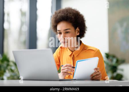 Fernschulung. Konzentrierte lateinische Dame, die sich einen Videokurs auf dem Laptop ansieht und Notizen macht, und virtuelle Lektion lernt Stockfoto