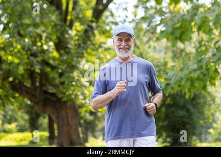 Älterer grauhaariger Mann, der im Park joggt, Rentner lächelt zufrieden im Park zwischen den Bäumen, trägt Sportkleidung und eine Kappe auf dem Kopf. Stockfoto