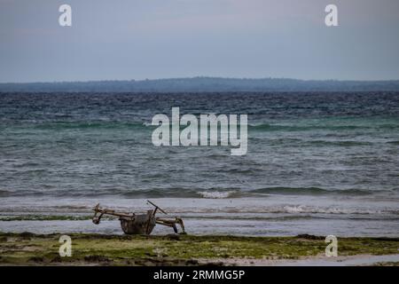 Ein kleines hölzernes Ausriggerkanu ruht am Ufer neben blauem Küstenwasser. Stockfoto