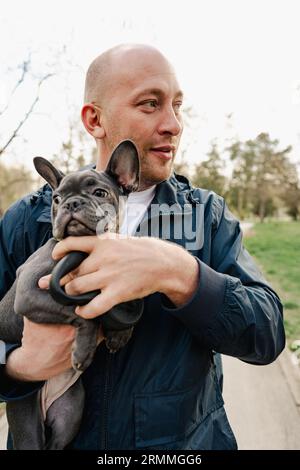 Mann, der seinen französischen Bulldoggen-Welpen in den Händen hält, während er im Park spaziert Stockfoto