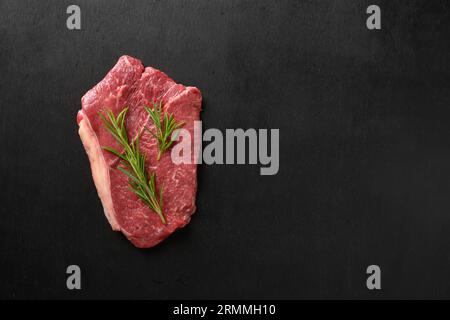 Rohes marmoriertes Ribeye-Steak mit Rosmarinzweig auf schwarzem Hintergrund. Draufsicht. Flat-Lay-Kopierbereich. Stockfoto