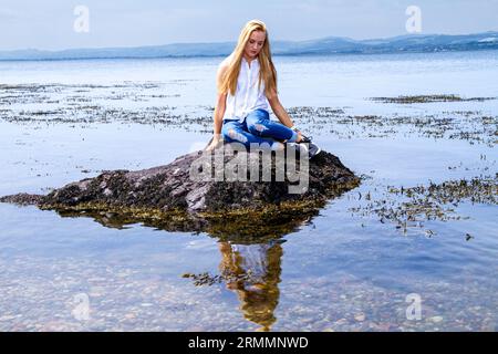 Eine wunderschöne blonde Frau, Rhianna Martin, sitzt auf den Felsen am Wormit Beach im Fife County, Schottland und genießt das angenehme Wetter, Großbritannien Stockfoto