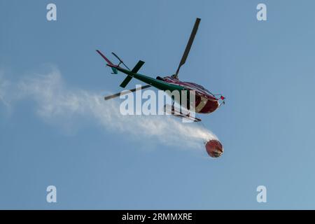 Blick von unten auf einen Hubschrauber, der einen Behälter mit Wasser trägt, um ein Feuer auszulösen Stockfoto