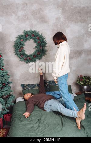 Glückliches Paar, das vor Weihnachten zu Hause genießt. Winterpullover für die ganze Familie auf dem Bett in der Nähe des Weihnachtsbaums mit Weihnachtsgeschenken. Leerzeichen kopieren Stockfoto
