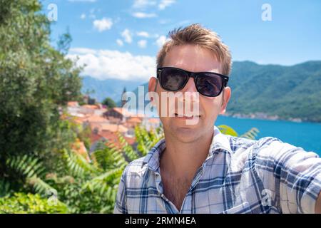 Glücklicher Mann mit Sonnenbrille, der Selfie im Freien in der Bucht von Kotor, Montenegro, nimmt. Männlicher Tourist, der Spaß im Sommerurlaub hat. Lächelnder Kerl, der Urlaub genießt Stockfoto
