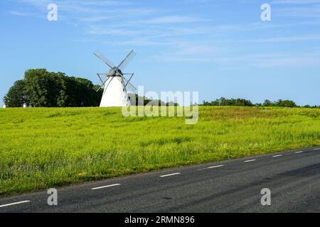 Historische weiße Windmühle auf einer grünen Wiese an der Seite der schwarzen Asphaltstraße, blauer Himmel Hintergrund Stockfoto