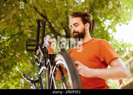 Aktiver und gesunder Radfahrer, der das Rad seines Fahrrads auf Schäden im Hof untersucht. Das Bild zeigt einen jungen kaukasier, der ein defektes Bauteil mit Schmiermittel fixiert und so eine sichere Fahrt gewährleistet Stockfoto