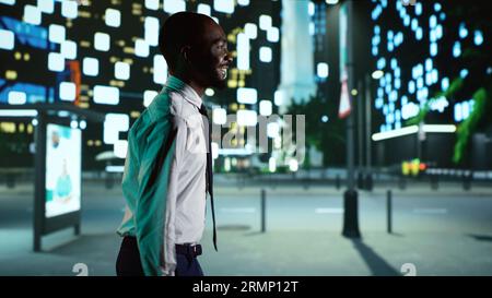 Der afroamerikanische Mann spaziert nachts in die Innenstadt und bewundert moderne Wolkenkratzer unter den Lichtern der Stadt. Fröhliche Person in trendigem Anzug zu Fuß in der Nähe des Geschäftsviertels im Stadtzentrum. Stockfoto