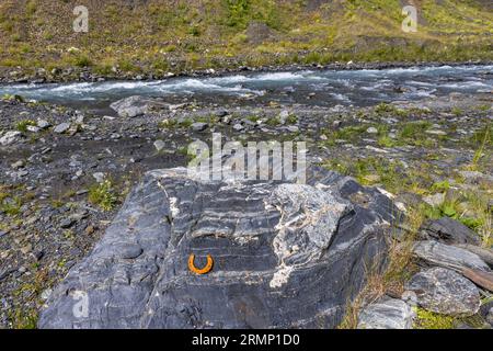 Ein rostiger Hufeisen liegt auf einem dunklen Felsbrocken mit hellen Adern am Ufer eines Gebirgsflusses auf dem Weg von den Kvakhidi-Wiesen zum Atsunta-Pass in der Gemeinde Akhmeta, Region Kachetien, Georgien. Hinter dem Felsen schäumt der Fluss durch ein breites Geröllbett. Gras und niedrige Pflanzen wachsen zwischen den Steinen am Ufer. Die Packpferde auf den Trekkingtouren werden etwa viermal im Jahr unterwegs mit neuen Hufeisen ausgestattet. Die Wiesen liegen auf einer Höhe von etwa 2.400 Metern im oberen Kvakhidistskali-Tal im Tusheti-Nationalpark. Der Weg von Girevi zum Atsunta-Pass führt durch dieses Va Stockfoto