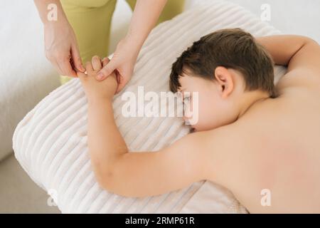 Von oben abgeschnittene Aufnahme einer nicht erkennbaren pädiatrischen Masseurin, die einem fünf Jahre alten Jungen, der auf dem Massagetisch liegt, Finger- und Palmenmassage macht Stockfoto