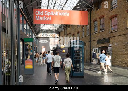 Leute, die durch den überdachten Old Spitalfields Markt gehen, mit einem großen Werbeschild oben und einer grünen Telefonbox. London, England Stockfoto