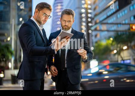 Geschäftsleute prüfen ihre E-Mails und lesen wichtige Dokumente auf einem Tablet vor dem Hintergrund einer Stadt bei Nacht. Zwei Geschäftsleute benutzen Tablets, um im Freien zu arbeiten Stockfoto
