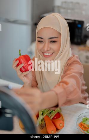 Eine attraktive asiatische muslimische Influencerin oder Food-Bloggerin zeichnet ihr Kochvideo in der Küche auf oder streamt ihr Essensrezept live auf Social Media Stockfoto