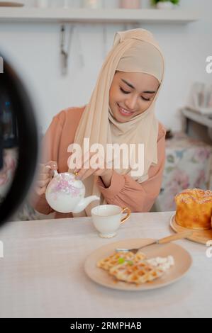 Eine schöne und glückliche asiatische Muslimin trinkt Nachmittagstee an einem Tisch in der Küche. Hausgemachtes Gebäck, Frühstück, Bäckerei Stockfoto