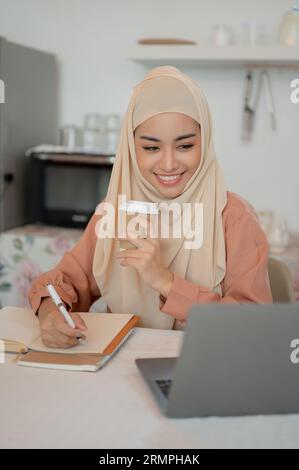 Eine wunderschöne asiatische muslimische Studentin studiert an einem Tisch in der Küche eine Online-Klasse und schreibt etwas in ein Buch, während sie Kaffee schlürfen Stockfoto
