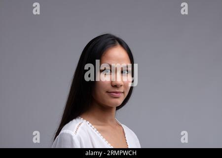 Attraktive dunkelhaarige Frau mit einem ruhigen Ausdruck, die sich mit einem ruhigen Lächeln auf grauem Hintergrund zur Kamera dreht Stockfoto