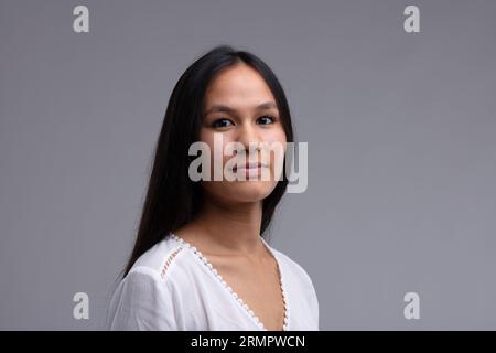 Schöne junge brünette Frau mit dicken schwarzen Haaren, die weiße Bluse trägt. Blick auf die Kamera mit schwachem Lächeln. Nahaufnahme eines Studioporträts auf grauem Hintergrund Stockfoto