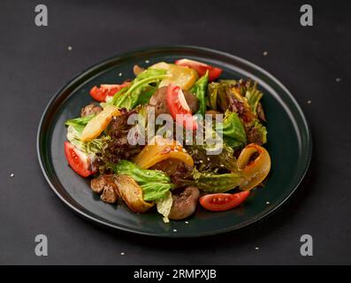 Warmer Salat mit knusprigen Salatblättern, gebratener Hühnerleber, karamellisierten Birnenscheiben, Rosinen und Balsamico-Sauce auf einem schwarzen Teller Stockfoto