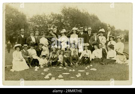 Postkarte einer Teeparty-Gruppe im Freien auf einem Feld irgendwo in Großbritannien - vielleicht ein Kirchenausflug, Tassen und Untertassen verstreut, eine erstaunliche Auswahl an Hüten und Outfits. Faszinierende Charaktere, Männer, Frauen und Kinder, sogar Babys. Im Hintergrund steht eine Kutsche mit einer mürrisch aussehenden älteren Frau, die einen Gesichtsschleier trägt, vielleicht um lästige Fliegen fernzuhalten. Foto um 1907, möglicherweise Newport Area, Wales, Großbritannien Stockfoto