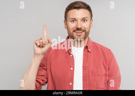 Glücklicher überraschter unrasierter Mann, der den Zeigefinger nach oben auf den freien Platz im Hintergrund zeigt und die Kamera betrachtet. Stockfoto