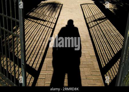 Voraussagender Schatten der Person stehend, Beine auseinander und Hände in Taschen, zwischen zwei halb offenen, schmiedeeisernen Toren Stockfoto