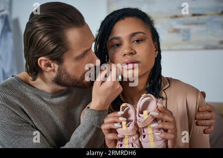 Fürsorglicher Mann, der die afroamerikanische Frau beruhigt, weint und Babykleidung hält, Fehlgeburtenkonzept Stockfoto