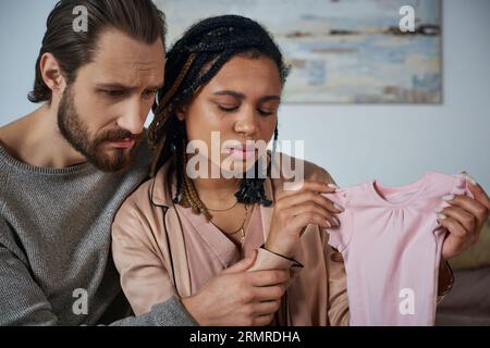 Trauriger Mann, der die afroamerikanische Frau beruhigt, weint und Babykleidung hält, Fehlgeburtenkonzept Stockfoto