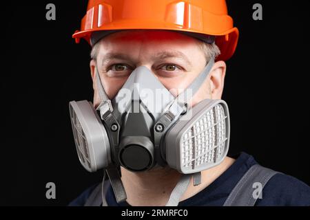 Ein Mann mit Schutzhelm und Atemschutzmaske zum Schutz vor Staub und Gasen auf schwarzem Hintergrund. Stockfoto