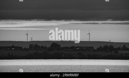 Windkraftanlagen, erneuerbare Energien, auf einem See bei Sonnenuntergang in Schwarz-weiß. Sauberer Strom für den Klimawandel. Bewölkter Himmel mit oranger Sonne in Scan Stockfoto