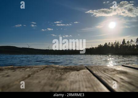 Blick über eine Fußgängerbrücke in das blaue Wasser eines Sees. Wald am Ufer des Sees. Die Sonne scheint zur Arbeitszeit. Windturbinen im Hintergrund Stockfoto