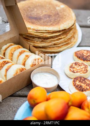 Hüttenkäse-Pfannkuchen, Syrniki in einer Papierkiste und stapeln dünne Pfannkuchen auf einem Holztisch. Lieferung gesundes und köstliches Frühstück am Morgen. Stockfoto