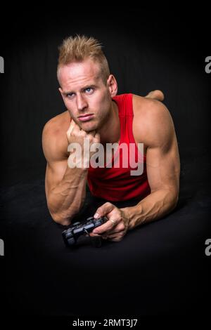 Ein Mann in einem roten Tank, der auf dem Boden lag, gelangweilt, mit einem Videospiel-Joystick in der Hand Stockfoto