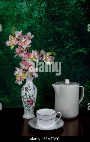 Künstliche Blumen in einer weiß verzierten Porzellanvase mit weißer Teekanne und Teetasse und Untertasse auf einer hölzernen Tischplatte mit meliertem Hintergrund Stockfoto