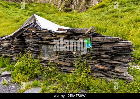 Eine niedrige Hütte aus trocken gestapelten Schieferplatten steht neben dem Pfad westlich von Chontio in der Gemeinde Akhmeta, Region Kachetien, Georgien. Ein graues Tarnnetz bedeckt das Dach. Eine grüne Plastikflasche hängt an einer Schnur von einem Holzstab über der Mauer, möglicherweise als einfacher Wasserspender. Eine große, glatte Steinplatte ist in die Mauer eingelassen. Kräuter wachsen vor der Mauer. Dahinter steigt ein grüner Hang zu einer Felswand an. Die Hütte wird möglicherweise von Hirten als Sommerquartier genutzt. Der Pfad von Girevi zum Atsunta-Pass folgt dem Kvakhidistskali-Tal und führt durch das verlassene Dorf Chontio. C Stockfoto