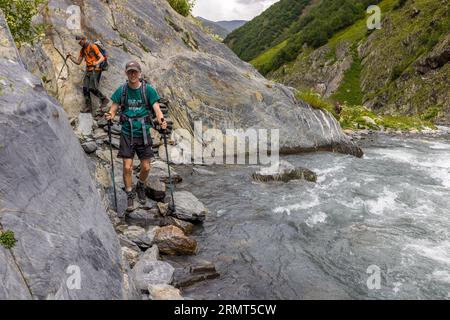 Ein junger Mann in einem grünen T-Shirt geht mit Trekkingstöcken über Steinplatten am Ufer eines rauschenden Gebirgsflusses westlich von Chontio in der Gemeinde Akhmeta, Region Kachetien, Georgien. Der Pfad verläuft am Fuße von glatten grauen Schieferfelsen direkt neben dem Wasser. Hinter ihm klettert ein Mann in einem orangefarbenen T-Shirt mit einem großen Rucksack über die Steine. Rechts schäumt der Fluss um einen Felsblock. Bewaldete und felsige Hänge erheben sich im Hintergrund. Der Weg von Girevi zum Atsunta-Pass folgt dem Kvakhidistskali-Tal und führt durch das verlassene Dorf Chontio. Chontio (Georgisch ჭონთიო) liegt Stockfoto