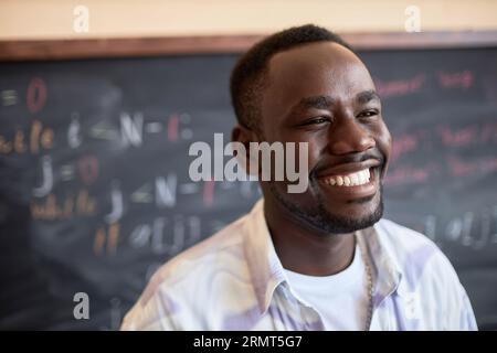Lachender afroamerikanischer Mann in weißer Casualwear, der vor der Kamera gegen Tafel steht, mit Formeln oder Gleichungen, die mit Kreide geschrieben sind Stockfoto