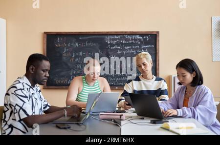 Vier junge interkulturelle Kollegen in Casualwear, die sich Bildschirme mit mobilen Gadgets ansehen, während sie einzeln an ihren neuen Projekten arbeiten Stockfoto