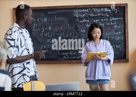 Asiatische Studentin schaut sich die Seite des offenen Buches an, während sie an der Tafel mit Formeln steht und vor Klassenkameraden Bericht erstattet Stockfoto
