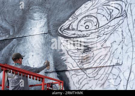 Southend on Sea, Essex, Großbritannien. 30. August 2023. Southend, das als Europas größtes Street-Art-Festival bezeichnet wird, feiert mit dem City Jam-Event seinen neuen Stadtstatus weiter. Über 200 lokale, nationale und internationale Straßenkünstler zeigen ihre Fähigkeiten an 100 Orten im Stadtzentrum. Einige provisorische Mauern wurden errichtet, aber die Künstler schmücken auch die Seiten von Gebäuden und andere dauerhafte Strukturen. Vom ersten bis zum dritten September haben die Künstler mit der Arbeit an den größeren Wandgemälden begonnen, um für die Veranstaltung bereit zu sein. Männlicher Künstler, der an grossen Fototapeten arbeitet Stockfoto