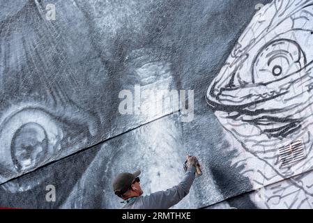 Southend on Sea, Essex, Großbritannien. 30. August 2023. Southend, das als Europas größtes Street-Art-Festival bezeichnet wird, feiert mit dem City Jam-Event seinen neuen Stadtstatus weiter. Über 200 lokale, nationale und internationale Straßenkünstler zeigen ihre Fähigkeiten an 100 Orten im Stadtzentrum. Einige provisorische Mauern wurden errichtet, aber die Künstler schmücken auch die Seiten von Gebäuden und andere dauerhafte Strukturen. Vom ersten bis zum dritten September haben die Künstler mit der Arbeit an den größeren Wandgemälden begonnen, um für die Veranstaltung bereit zu sein. Männlicher Künstler, der an grossen Fototapeten arbeitet Stockfoto