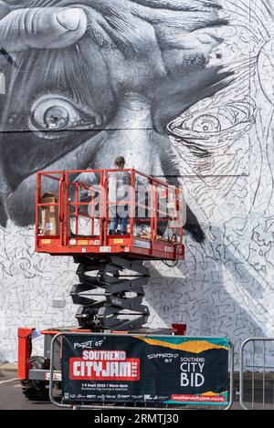 Southend on Sea, Essex, Großbritannien. 30. August 2023. Southend, das als Europas größtes Street-Art-Festival bezeichnet wird, feiert mit dem City Jam-Event seinen neuen Stadtstatus weiter. Über 200 lokale, nationale und internationale Straßenkünstler zeigen ihre Fähigkeiten an 100 Orten im Stadtzentrum. Einige provisorische Mauern wurden errichtet, aber die Künstler schmücken auch die Seiten von Gebäuden und andere dauerhafte Strukturen. Vom ersten bis zum dritten September haben die Künstler mit der Arbeit an den größeren Wandgemälden begonnen, um für die Veranstaltung bereit zu sein. Männlicher Künstler, der an grossen Fototapeten arbeitet Stockfoto