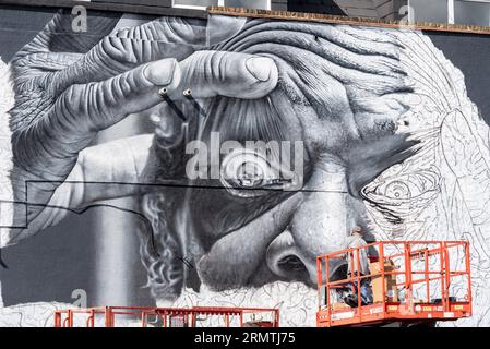 Southend on Sea, Essex, Großbritannien. 30. August 2023. Southend, das als Europas größtes Street-Art-Festival bezeichnet wird, feiert mit dem City Jam-Event seinen neuen Stadtstatus weiter. Über 200 lokale, nationale und internationale Straßenkünstler zeigen ihre Fähigkeiten an 100 Orten im Stadtzentrum. Einige provisorische Mauern wurden errichtet, aber die Künstler schmücken auch die Seiten von Gebäuden und andere dauerhafte Strukturen. Vom ersten bis zum dritten September haben die Künstler mit der Arbeit an den größeren Wandgemälden begonnen, um für die Veranstaltung bereit zu sein. Männlicher Künstler, der an grossen Fototapeten arbeitet Stockfoto