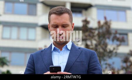 Hübscher kaukasischer, stilvoller Mann mit ernstem Gesicht, der in der Stadt auf der Straße steht und per Smartphone SMS schreibt. Junger erfolgreicher Geschäftsmann, der Anzug schreibt Stockfoto