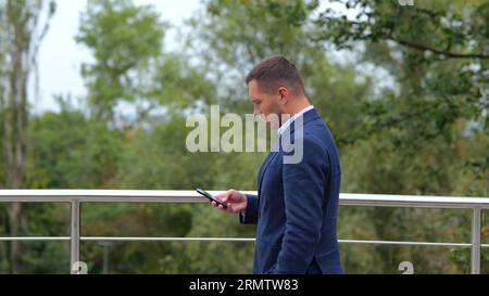 Seitenansicht des jungen Kaukasier, der Anzug trägt, der auf der Brücke in der Stadt draußen spaziert und auf dem Smartphone SMS schreibt. Zeitlupe. Männlicher Unternehmer läuft Stockfoto