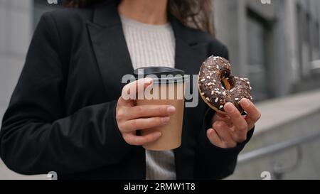 Junger Kaukasier mit braunem Haar, der schwarze Jacke trägt, Kaffee trinkt und SchokoladenDonut zwischen Wolkenkratzern isst. Im Freien, Snacks. Stockfoto