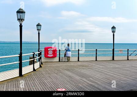 Eine einsame weibliche Figur, die am Ende des Worthing Pier an einem klaren Sommertag in Worthing West Sussex England auf das Meer blickt Stockfoto