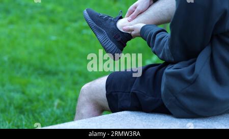 Seitenansicht junger kaukasischer Mann, der auf einer Betonbank sitzt und sein Bein nach dem Workout verletzt. Im Freien. Sportler im Black-Spot-Outfit entspannend, massierend Stockfoto