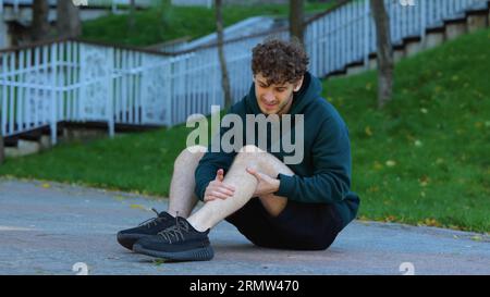 Sportler sitzt auf dem Boden und dehnt Knie, Bein und Muskeln im grünen Park vor Outdoor-Aktivitäten. Mann, der vorher sein Knie und seine Muskeln massiert Stockfoto