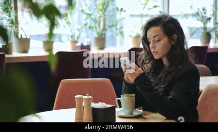 Nahaufnahme, Zeitlupe. Kaukasische Frau sitzt im Café, trinkt heißen Tee mit Pfefferminz, scrollt und schreibt auf ihrem Telefon. Stockfoto